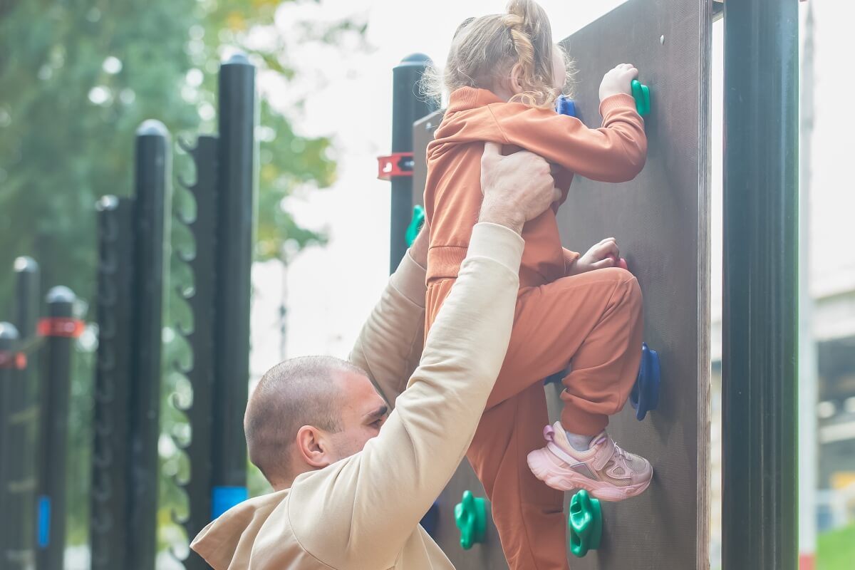Dziecko wspina się z pomocą na ściankę wspinaczkową ogrodową ze sklejki.